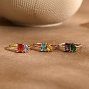 May include: Three rings with colorful gemstones are displayed. The rings feature rectangular and pear-shaped stones in red, purple, blue, yellow, green, and purple. The rings are set on a brown surface.