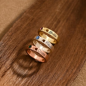 Op de afbeelding: Vier gepersonaliseerde ringen in goud, zilver en roségoud, elk met een andere edelsteen en gegraveerde naam. De ringen zijn gestapeld op een houten oppervlak, wat de op maat gemaakte sieraden laat zien.