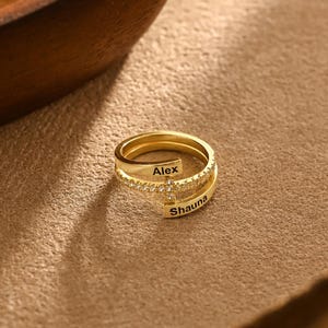 Puede incluir: Un anillo dorado con los nombres "Alex" y "Shauna" grabados. El anillo tiene un diseño de doble banda, con una banda adornada con pequeñas piedras preciosas transparentes. El anillo se muestra sobre una superficie marrón.
