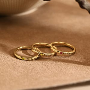 Può includere: Tre anelli d'oro con testo inciso e piccole pietre preziose. Un anello ha una gemma verde e legge "Nicole 10.08". Un altro ha una gemma gialla e legge "I love you". Il terzo anello ha una gemma rossa e legge "I'm listening".
