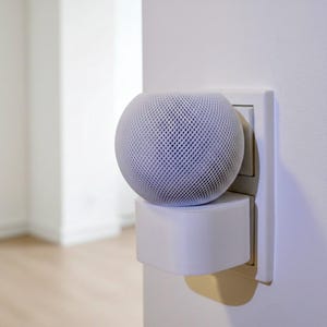 Könnte beinhalten: Ein weißer, kugelförmiger Smart-Lautsprecher mit einem Mesh-Außenmaterial ist in eine weiße Steckdose eingesteckt. Der Lautsprecher sitzt auf einem weißen Netzteil und erzeugt eine minimalistische Ästhetik. Der Hintergrund ist ein weicher, verschwommener Innenraum.