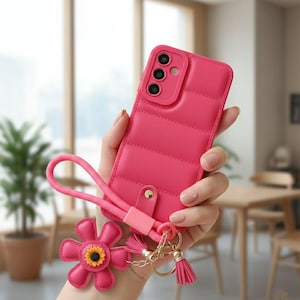 Puede incluir: Funda de teléfono rosa brillante con diseño acolchado. Incluye una correa de muñeca a juego y un charm en forma de flor con centro amarillo. Una pequeña borla rosa cuelga del llavero de la funda.