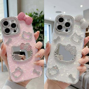 Peut inclure: Deux coques de t&eacute;l&eacute;phone, une rose et une blanche, chacune orn&eacute;e de d&eacute;corations scintillantes. Les coques pr&eacute;sentent un motif Hello Kitty avec un n&oelig;ud et d'autres &eacute;l&eacute;ments d&eacute;coratifs. L'objectif de la cam&eacute;ra est entour&eacute; d'un cadre argent&eacute;.