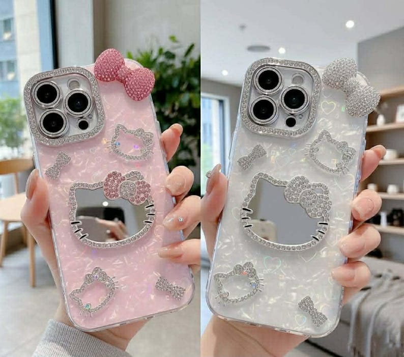 Peut inclure: Deux coques de t&eacute;l&eacute;phone, une rose et une blanche, chacune orn&eacute;e de d&eacute;corations scintillantes. Les coques pr&eacute;sentent un motif Hello Kitty avec un n&oelig;ud et d'autres &eacute;l&eacute;ments d&eacute;coratifs. L'objectif de la cam&eacute;ra est entour&eacute; d'un cadre argent&eacute;.