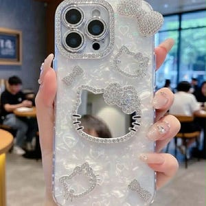 Peut inclure: Coque de t&eacute;l&eacute;phone transparente avec un fond nacr&eacute; blanc, orn&eacute;e de strass &eacute;tincelants. La coque pr&eacute;sente un motif Hello Kitty, des n&oelig;uds et des c&oelig;urs. L'objectif de l'appareil photo est entour&eacute; d'une bordure en strass.