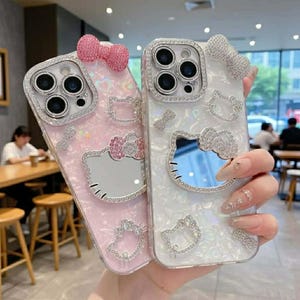 Peut inclure: Deux coques de t&eacute;l&eacute;phone d&eacute;coratives. L'une est rose avec un n&oelig;ud et un motif Hello Kitty, l'autre est blanche avec un n&oelig;ud et un motif Hello Kitty. Les deux coques sont orn&eacute;es de strass autour de l'appareil photo et du motif Hello Kitty.