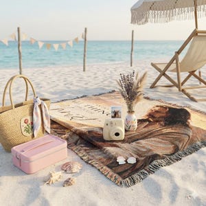 Puede incluir: Una escena de picnic en la playa con un bolso tote tejido, una fiambrera rosa y una manta con una imagen religiosa. También hay una cámara instantánea beige, un jarrón con flores secas y conchas marinas. Una sombrilla y una silla de playa están en el fondo.
