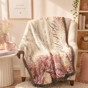 Puede incluir: Una manta tejida de color crema drapeada sobre una silla. La manta presenta las palabras "Best Mom" y un diseño floral en tonos rosas y crema. Los bordes están acabados con un ribete con flecos.