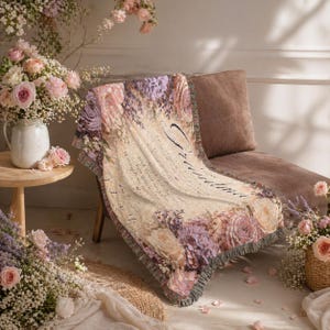 Puede incluir: Una manta floral con fondo crema y diseño de rosas y lavanda en tonos rosas y morados. La manta tiene un borde con flecos y texto. La manta está sobre una silla marrón.