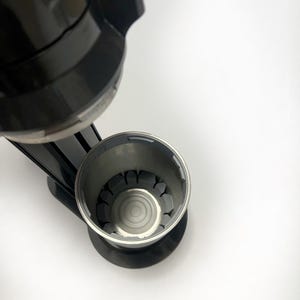 Puede incluir: Una cafetera negra con una cesta de filtro de acero inoxidable. La cesta del filtro tiene una serie de soportes grises y acanalados. La cafetera está colocada sobre un fondo blanco.