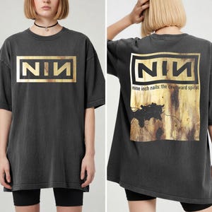 May include: Dark grey t-shirt with a gold rectangular logo reading "NIN". The back of the shirt displays the same logo and the text "nine inch nails: the downward spiral" with a distressed graphic.