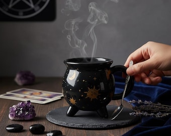 May include: A black ceramic cauldron-shaped mug with a handle and three feet. The mug is decorated with gold moons, stars, and suns. Steam rises from the mug, suggesting a hot beverage. A silver spoon rests on the handle. The mug sits on a black slate coaster.