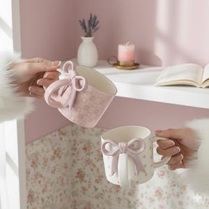 Puede incluir: Dos tazas de cerámica rosa y blanca con asas y detalles en forma de lazo. Una taza presenta un patrón de cuadros con corazones, mientras que la otra tiene un diseño de lunares. Las tazas están sobre un fondo rosa suave.