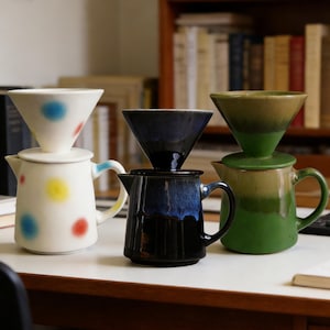 Puede incluir: Una colección de cafeteras de cerámica para verter en varios colores. Una es blanca con lunares de colores, otra es azul oscuro y la tercera es verde y marrón. Cada una tiene un filtro en forma de cono a juego.