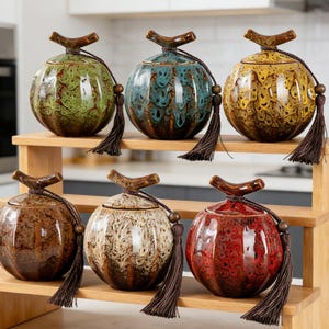 May include: Six decorative ceramic jars in various colors, including green, blue, and yellow, with textured surfaces and wooden-look handles. Each jar has a tassel and sits on a wooden shelf. The jars are round and have a lid.