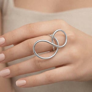 Könnte beinhalten: Ein silberfarbener Ring mit einem einzigartigen Unendlichkeits-Loop-Design. Der Ring wird an einer Hand mit einer neutral getönten Maniküre getragen. Das Design des Rings zeigt zwei ineinander verschlungene Schlaufen, die eine moderne und minimalistische Ästhetik schaffen.