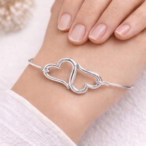 Könnte beinhalten: Ein silbernes Armreif-Armband mit zwei ineinandergreifenden Herzformen. Das Armband besteht aus dünnem, flexiblem Metall und wird am Handgelenk getragen. Die Herzen sind verbunden und bilden ein durchgehendes Design, das Liebe und Verbundenheit symbolisiert.