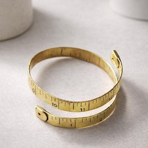 Puede incluir: Un brazalete dorado diseñado para parecerse a una cinta métrica. El brazalete está enrollado, con números y marcas similares a los de una cinta métrica. Un accesorio único.