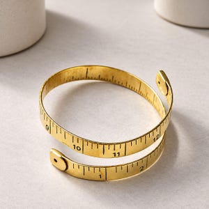May include: A gold-toned metal bracelet designed to resemble a flexible measuring tape. The band features black engraved markings and numbers ranging from 1 to 12, with a coiled, overlapping structure and flat, rounded ends at each terminal point.