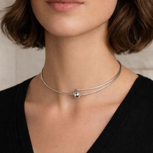 Könnte beinhalten: Ein silbernes Choker-Halsband mit minimalistischem Design. Die Halskette hat zwei parallele, dünne Bänder, die sich um den Hals legen, verbunden durch eine kleine, runde, silberne Kugel. Die Halskette wird über einem schwarzen Oberteil getragen.