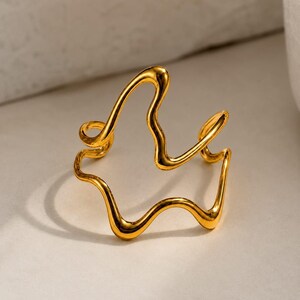 Può includere: Un anello color oro caratterizzato da un design a filo aperto, irregolare e ondulato. La fascia metallica si curva in molteplici anelli e punte fluide e organiche, creando una forma astratta continua con una finitura superficiale lucida.