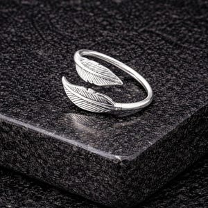 May include: A silver-colored ring with a leaf design. The ring features two detailed leaf shapes at the front, with a smooth, curved band that wraps around the finger. The ring is set against a dark, textured surface.