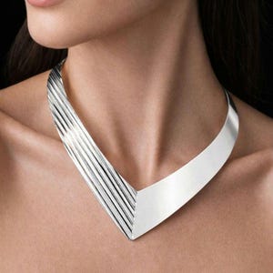 May include: A silver necklace with a V-shaped design. The left side has parallel, textured lines, and the right side is a smooth panel. The necklace is worn around the neck.