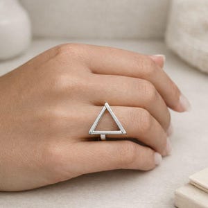 Puede incluir: Un anillo plateado con un diseño geométrico de triángulo. El anillo es de estilo minimalista, con forma de triángulo hueco. El anillo se muestra en una mano, mostrando su estética moderna y sencilla.