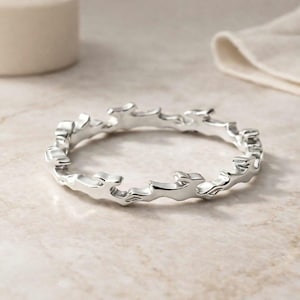 Puede incluir: Un anillo plateado con un diseño único y fluido. La superficie del anillo presenta un patrón orgánico en forma de ola, creando una apariencia texturizada. El anillo se muestra sobre una superficie de color claro.