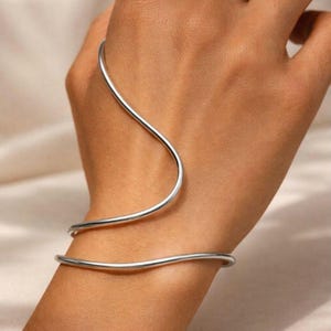 May include: A silver arm cuff bracelet. The jewellery features a minimalist design with two curved bands that wrap around the wrist and forearm. The bracelet is made of a shiny, smooth metal.