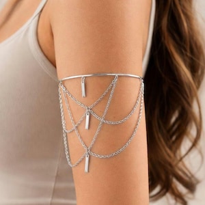 May include: Silver arm cuff with a delicate design. The cuff has a band that goes around the upper arm, with multiple chains cascading down. Rectangular charms are suspended from the chains. The jewelry is worn on the arm.