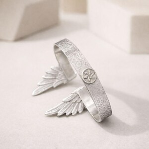 May include: A silver-toned bracelet with a textured band and wing-shaped accents. The bracelet features a small, circular emblem with a leaf design. The wings are detailed with individual feather lines, creating a delicate and artistic look.