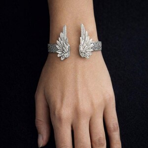 May include: Silver-toned cuff bracelet featuring an open design with detailed wing accents. The wings are positioned on either side of the wrist, with intricate feather detailing. The bracelet has a textured band and is displayed on a hand against a dark background.