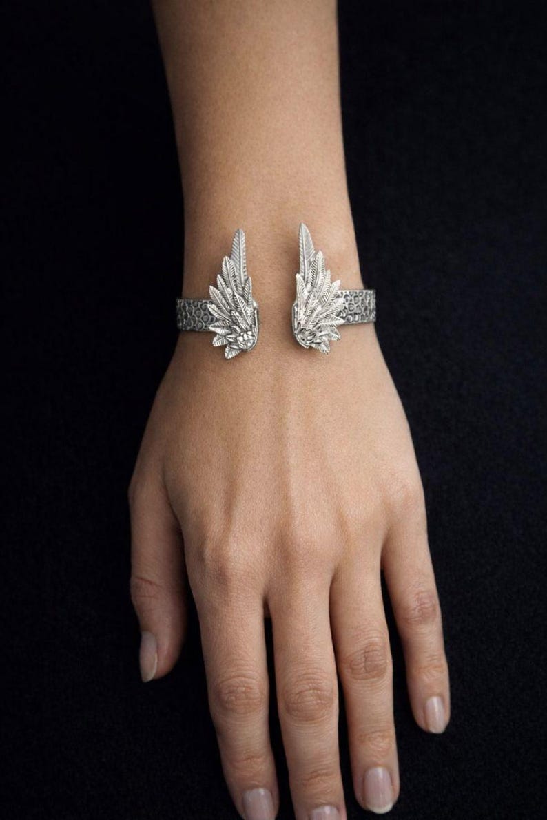 May include: Silver-toned cuff bracelet featuring an open design with detailed wing accents. The wings are positioned on either side of the wrist, with intricate feather detailing. The bracelet has a textured band and is displayed on a hand against a dark background.
