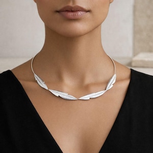 Può includere: Una collana d'argento con una serie di ciondoli a forma di foglie stilizzate. La collana è indossata su un top nero, con le foglie disposte in un design curvo. Il gioiello è un accessorio minimalista.