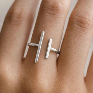 Op de afbeelding: Een zilverkleurige ring met een modern, minimalistisch ontwerp. De ring heeft twee verticale staven, de ene iets hoger dan de andere, verbonden door een band die de vinger omsluit. De ring wordt aan een vinger gedragen.