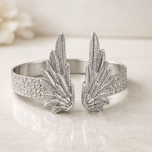 May include: Silver-toned cuff bracelet featuring a detailed feather wing design on each end. The bracelet has a textured band with a honeycomb pattern. A stylish accessory for everyday wear.
