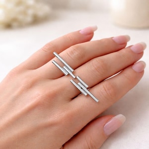 Puede incluir: Un anillo plateado con un diseño moderno. El anillo presenta múltiples barras rectangulares dispuestas de forma lineal. La joya se muestra en una mano con uñas cuidadas.