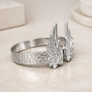 May include: A silver-toned bracelet with a textured band and two detailed wing accents. The wings are positioned upright, adding a unique, decorative element to the jewelry. The bracelet's design is intricate and eye-catching.