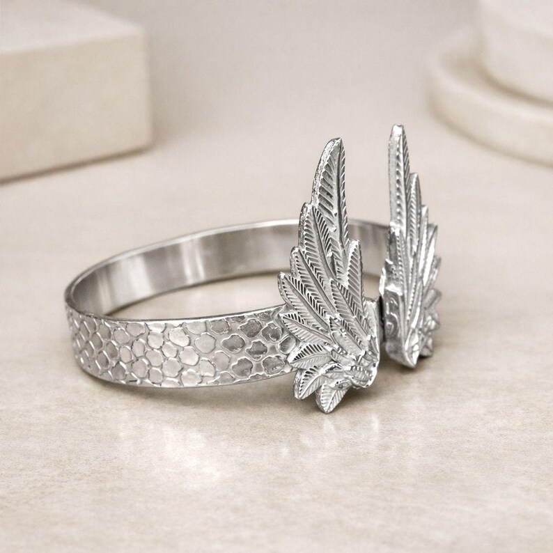 May include: A silver-toned bracelet with a textured band and two detailed wing accents. The wings are positioned upright, adding a unique, decorative element to the jewelry. The bracelet's design is intricate and eye-catching.