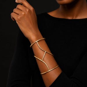 May include: A wide gold-toned metal cuff bracelet featuring a structural design with two horizontal bands connected by a central vertical bar and two diagonal bars, creating an open, geometric framework that wraps around the forearm.