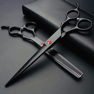 Puede incluir: Tijeras de peluquería negras con detalles rojos y un peine negro. Las tijeras están cruzadas sobre el peine, con un estuche de cuero negro en el fondo. Las tijeras tienen un mango curvo y una hoja afilada.