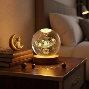 Puede incluir: Una escena decorativa de mesita de noche con una lámpara esférica de cristal brillante con un diseño de sistema solar, colocada sobre una base de madera. Un astronauta dorado en una luna creciente, libros y esferas decorativas completan la exhibición. La lámpara tiene unos 15 cm de diámetro.