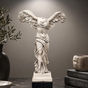 Puede incluir: Una estatua blanca con acabado desgastado de una figura alada sin cabeza sobre una base rectangular. La escultura presenta pliegues de tela fluidos e intrincados en el torso y las piernas, con grandes alas detalladas extendidas hacia los lados.