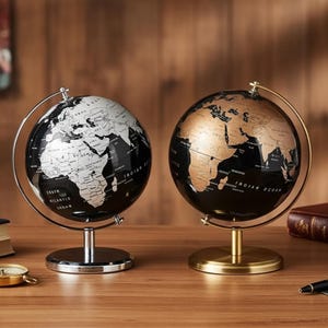 May include: Two decorative globes, one black and white, the other black and gold, are displayed on a wooden surface. The globes are mounted on metal stands. The black and white globe has a silver stand, the other a gold stand.