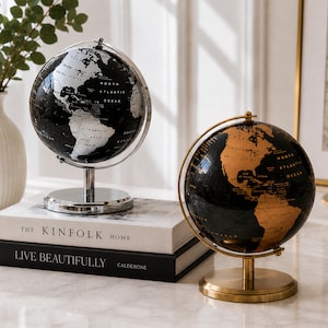 Può includere: Due mappamondi decorativi, uno nero e argento, l'altro nero e oro, su una superficie bianca. Il mappamondo nero e argento ha un supporto argentato, e quello nero e oro ha un supporto dorato. I testi "THE KINFOLK HOME" e "LIVE BEAUTIFULLY" sono visibili.
