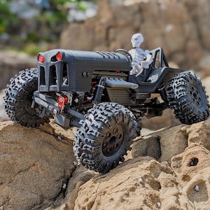 Może przedstawiać: Czarny, miniaturowy pojazd terenowy z dużymi, wytrzymałymi oponami i kierowcą-szkieletem. Pojazd ma czerwone akcenty z przodu i jest umieszczony na skalistej powierzchni, sugerując zabawę lub wystawę na świeżym powietrzu.