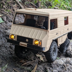 以下が含まれることがあります： 黒いタイヤと黄色のヘッドライトを備えた、ベージュ色の玩具トラック。トラックは、フロントグリルとフロントガラスを通して見える運転席を備えた箱型のデザインです。トラックは泥だらけの表面にあります。