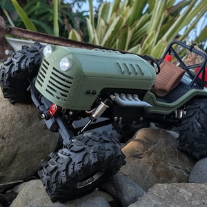 May include: A green and black toy off-road vehicle with large, black, knobby tires. The vehicle has a detailed engine compartment, headlights, and a roll cage. It is positioned on a rocky surface, suggesting an outdoor setting.