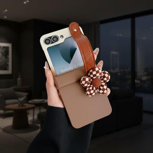 Puede incluir: Funda de teléfono beige y marrón para Galaxy Z Flip. Cuenta con una correa de cuero marrón y una flor decorativa con un diseño a cuadros. La pantalla del teléfono muestra "Galaxy Z Flip".